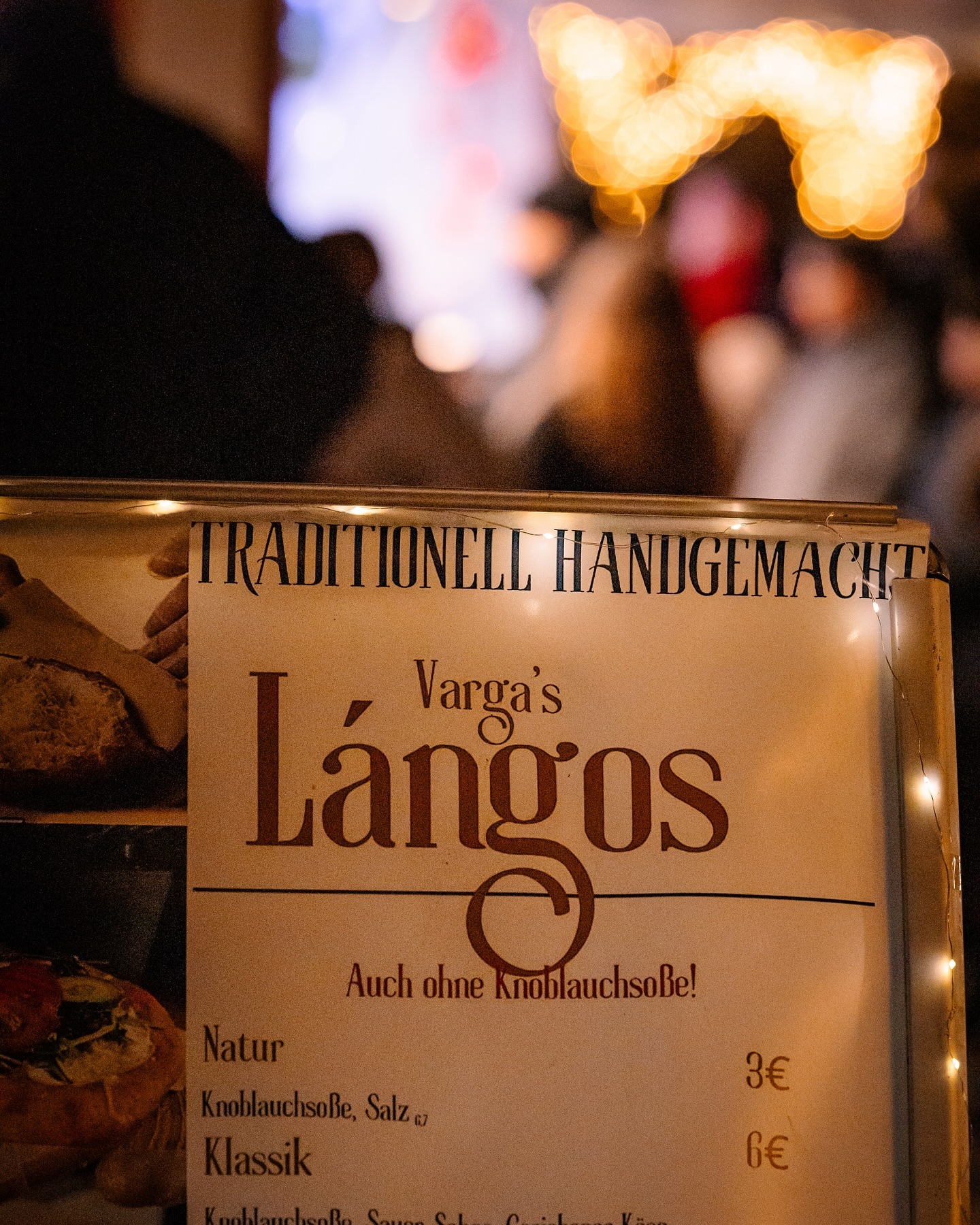 Ein Schild wirbt für traditionell handgemachte Lángos in verschiedenen Variationen mit Preisen. Im Hintergrund sind unscharf Menschen und Lichter zu sehen.