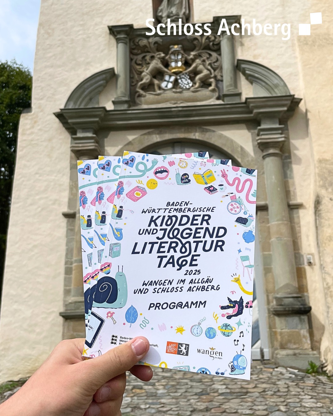 Ein farbiges Plakat wird vor einem steinernen Torbogen gehalten. Das Plakat bewirbt eine Veranstaltung für Kinderliteratur im Jahr 2025.