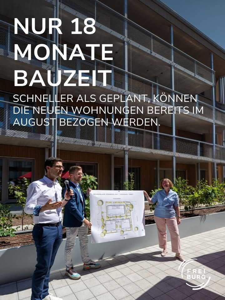 Drei Personen stehen vor einem modernen Wohngebäude und halten gemeinsam einen Grundriss, während eine Person ein Mikrofon hält. Der eingeblendete Text lautet: „NUR 18 MONATE BAUZEIT SCHNELLER ALS GEPLANT, KÖNNEN DIE NEUEN WOHNUNGEN BEREITS IM AUGUST BEZOGEN WERDEN.“ Unten rechts ist das Logo „FREI BURG“ zu sehen. (KI-Alternativtext)