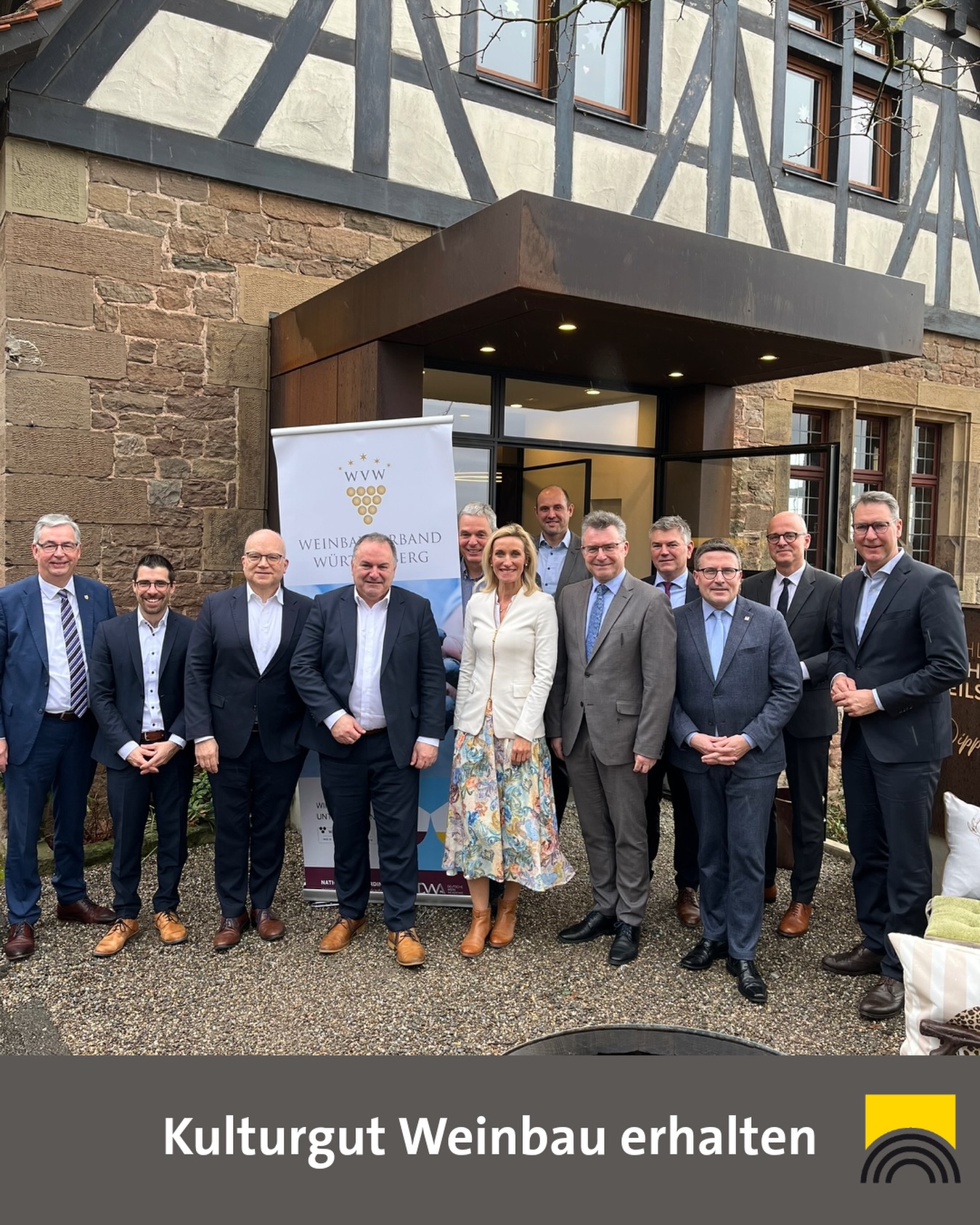 Dieser Alternativtext wurde KI-generiert: Eine Gruppe von Personen steht lächelnd vor einem Gebäude mit einem Banner und einer Steinmauer. Im Vordergrund befindet sich ein Banner mit Traubenmotiven und Text.