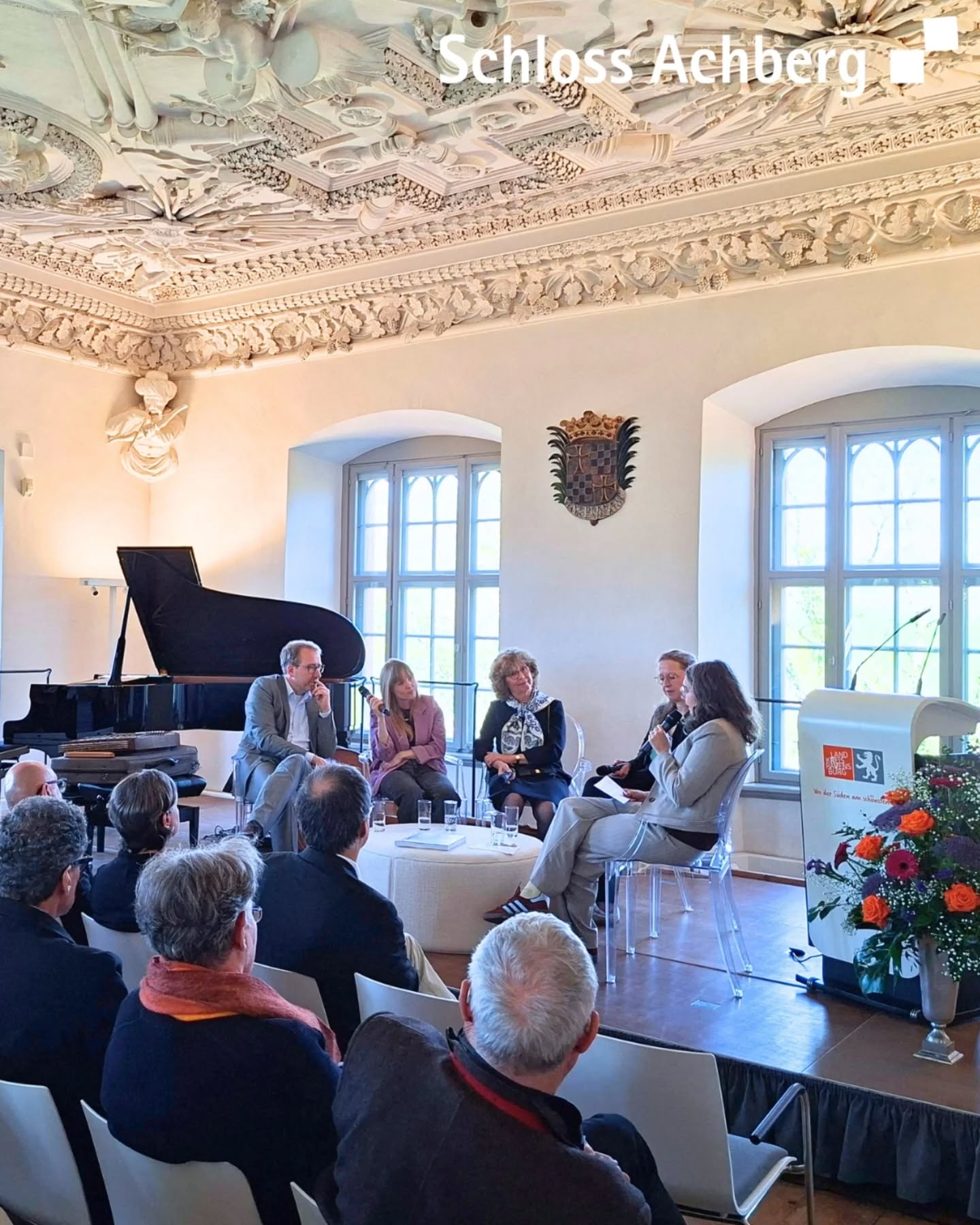 Dieser Alternativtext wurde KI-generiert: Innenaufnahme eines Raumes mit einer Gruppe von Menschen, die um einen Tisch sitzen und sich unterhalten. Im Hintergrund sind Fenster und ein Flügel zu sehen.