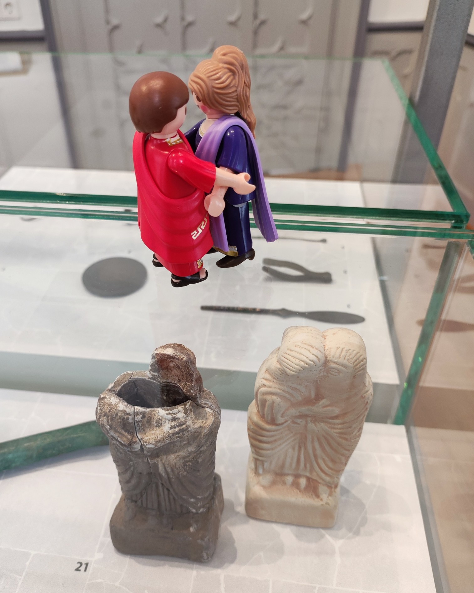 Dieser Alternativtext wurde KI-generiert: Zwei Plastikfiguren stehen auf einer Glasplatte über archäologischen Fundstücken. Die Szene ist in einem Ausstellungsraum zu sehen.