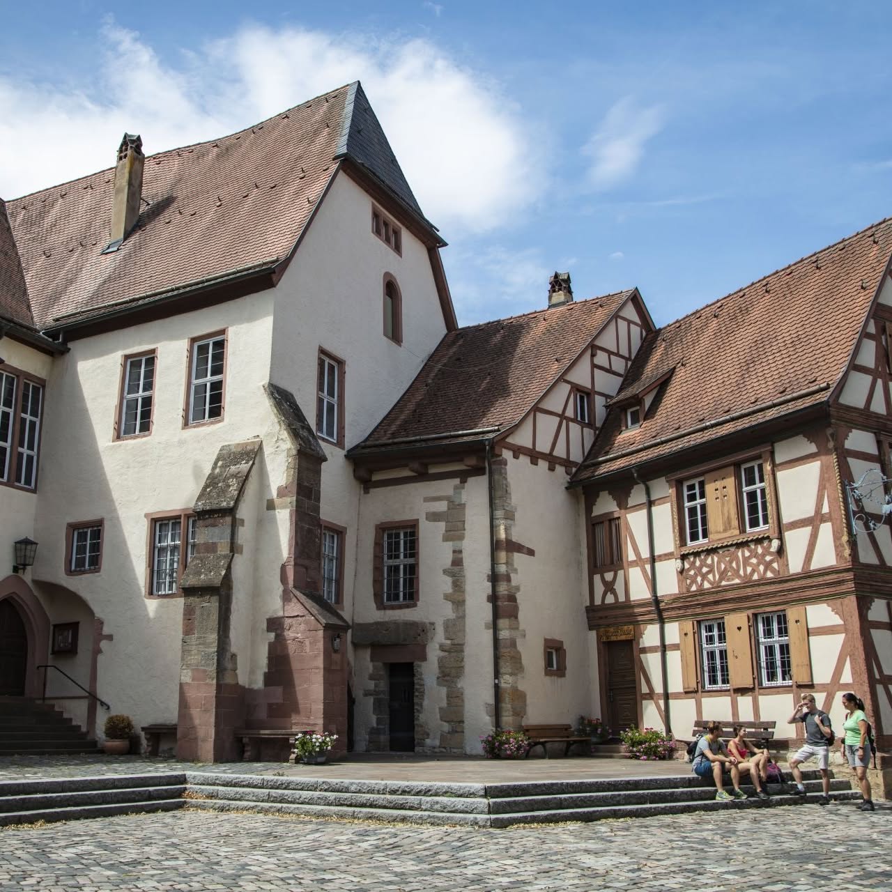 Ein mittelalterlicher Gebäudekomplex mit Fachwerk und Naturstein an einem sonnigen Tag. Mehrere Personen sitzen auf den Stufen vor dem Gebäude.