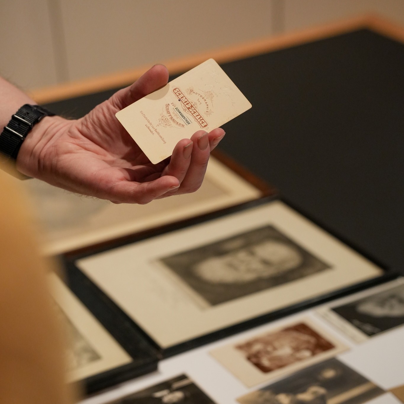 Eine Hand mit einer schwarzen Armbanduhr hält eine kleine, cremefarbene Karte, auf der in roter Schrift "PHOTOGRAPHIE VON SCHMID-SEILER ZU SCHWARZENBERG GRAF FLEISCHMANN" steht. Im unscharfen Hintergrund liegen auf einer schwarzen Fläche mehrere gerahmte und lose historische Fotografien. (KI-Alternativtext)