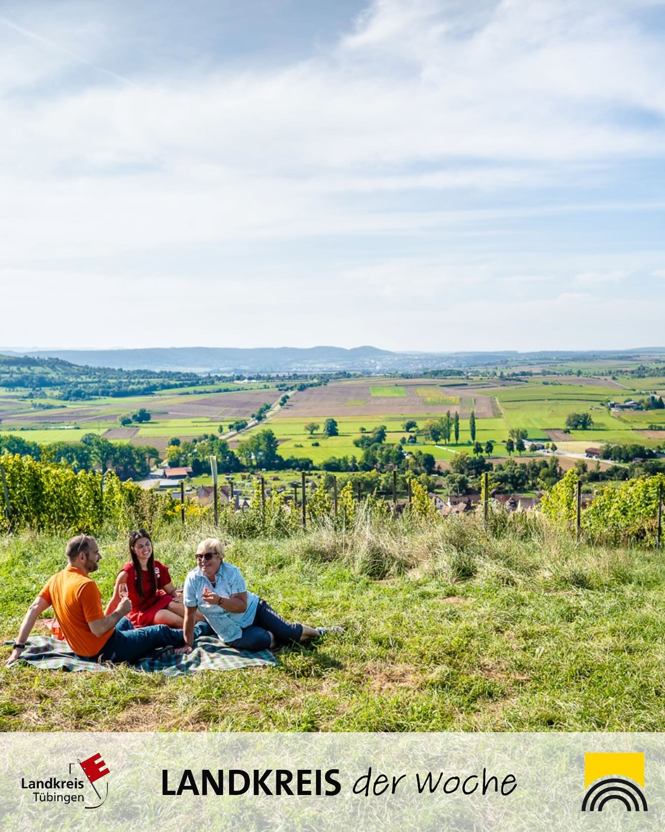 Eine farbige Aufnahme zeigt eine ländliche Landschaft mit Feldern und Hügeln. Im Vordergrund sitzen Personen auf einer Decke und scheinen die Aussicht zu genießen.