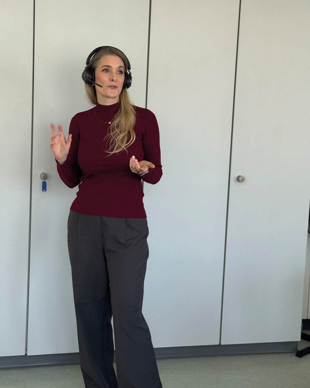 Dieser Alternativtext wurde KI-generiert: Eine Person mit langen Haaren trägt ein dunkelrotes Oberteil und eine Kopfhörer. Sie steht vor einer Tür und macht eine Geste mit der Hand.