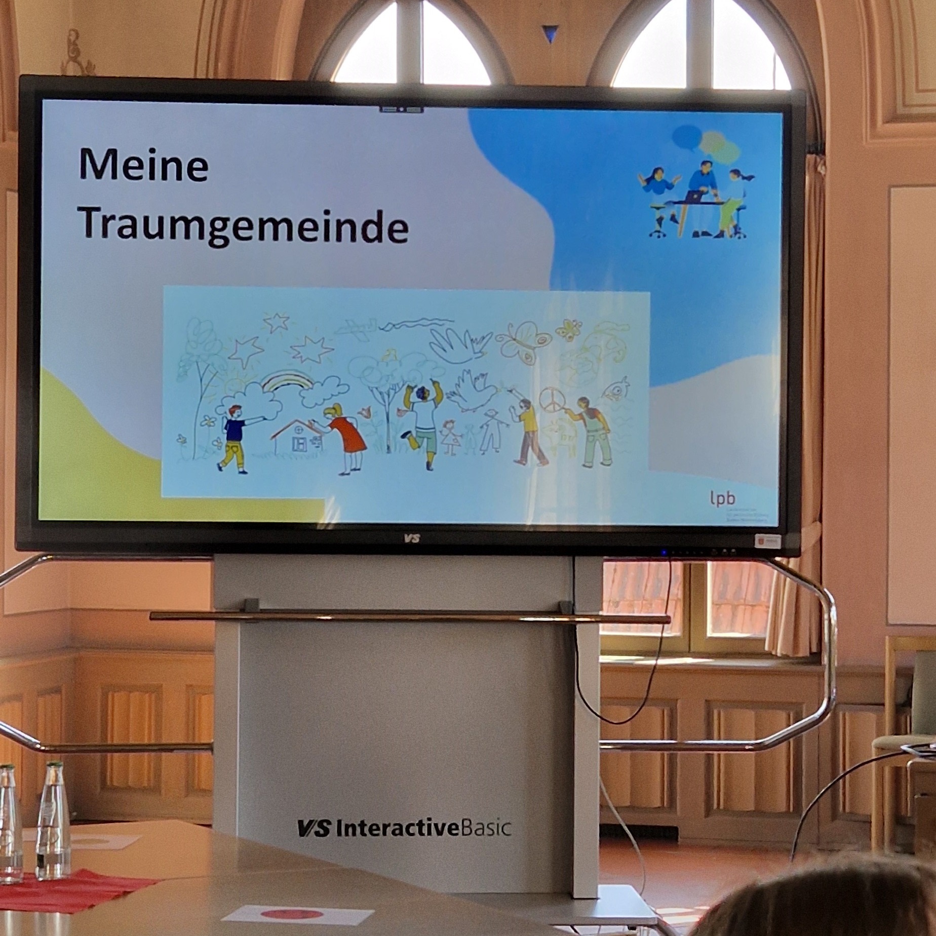Präsentationsbildschirm mit dem Titel „Meine Traumgemeinde“ und einer Zeichnung von Menschen, die gemeinsam malen, sowie Symbolen wie Regenbogen, Tauben und Friedenszeichen. Unten rechts steht „lpb“. Der Bildschirm steht auf einem Podest mit der Aufschrift „VS InteractiveBasic“. (KI-Alternativtext)