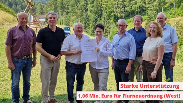 Acht Personen stehen im Freien auf einer Wiese vor einem bewaldeten Hang. Zwei Personen in der Mitte halten einen großen Scheck mit der Aufschrift „Optima (West)“ und dem Betrag 1.062.831,12 Euro. Ein roter Text unten rechts lautet: „Starke Unterstützung 1,06 Mio. Euro für Flurneuordnung (West)“. (KI-Alternativtext)