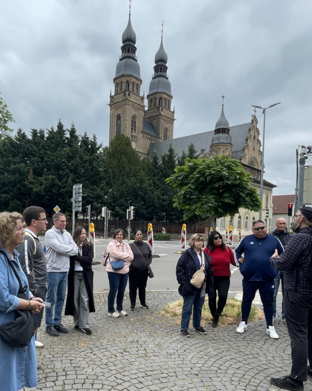 Gruppe von Menschen auf einem Kopfsteinpflaster vor einer Kirche mit zwei hohen Türmen unter bewölktem Himmel. Eine Person gestikuliert, als würde sie etwas erklären. (KI-Alternativtext)