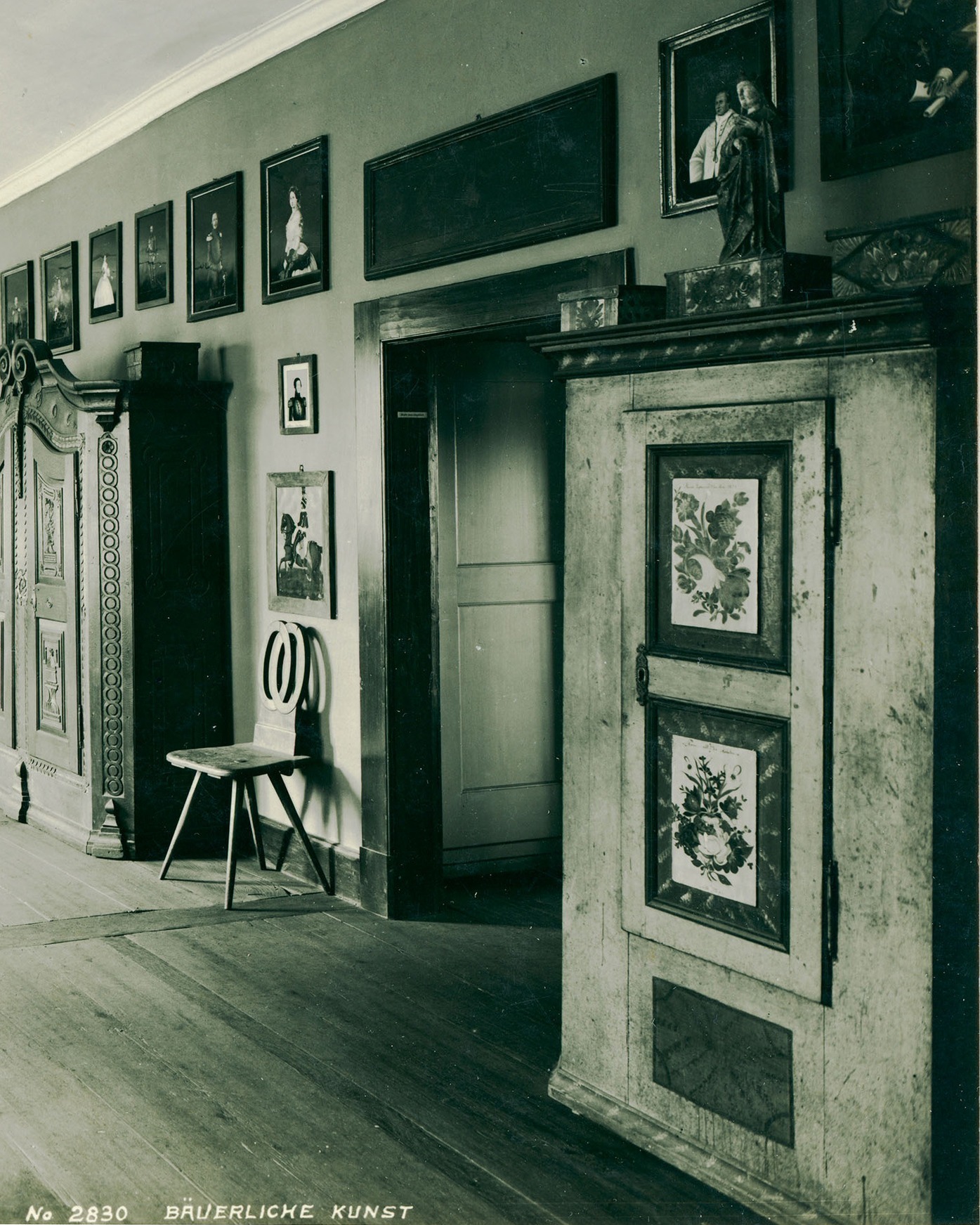 Dieser Alternativtext wurde KI-generiert: Schwarz-weißes Foto einer Innenraumansicht mit einer Galerie von Porträts an der Wand. Möbelstücke, darunter ein Schrank und ein Stuhl, stehen im Raum.