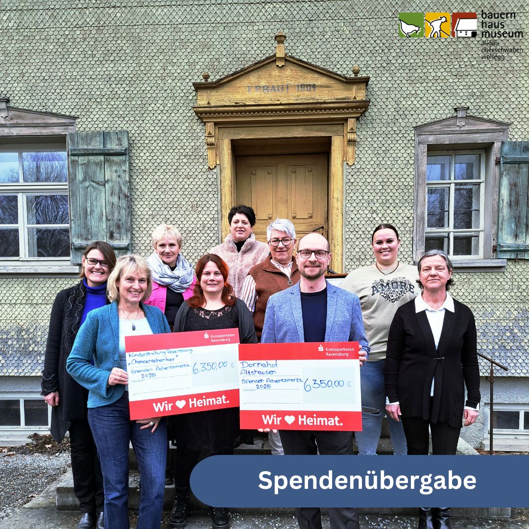 Dieser Alternativtext wurde KI-generiert: Eine Gruppe von Personen steht vor einem Gebäude mit einem Fenster und einer Tür. Sie halten Schilder mit Zahlen und Text, die eine Spendenübergabe darstellen.