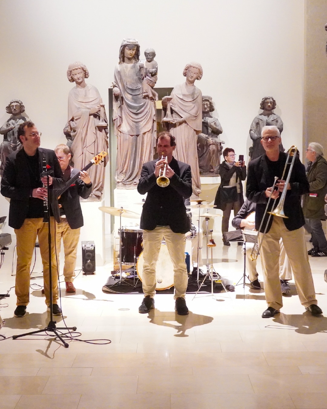 Dieser Alternativtext wurde KI-generiert: Eine Gruppe von Personen spielt Musikinstrumente in einem Innenraum vor mehreren steinernen Figuren. Das Bild ist farbig und zeigt ein Ensemble mit Trompeten, Gitarren und anderen Instrumenten.