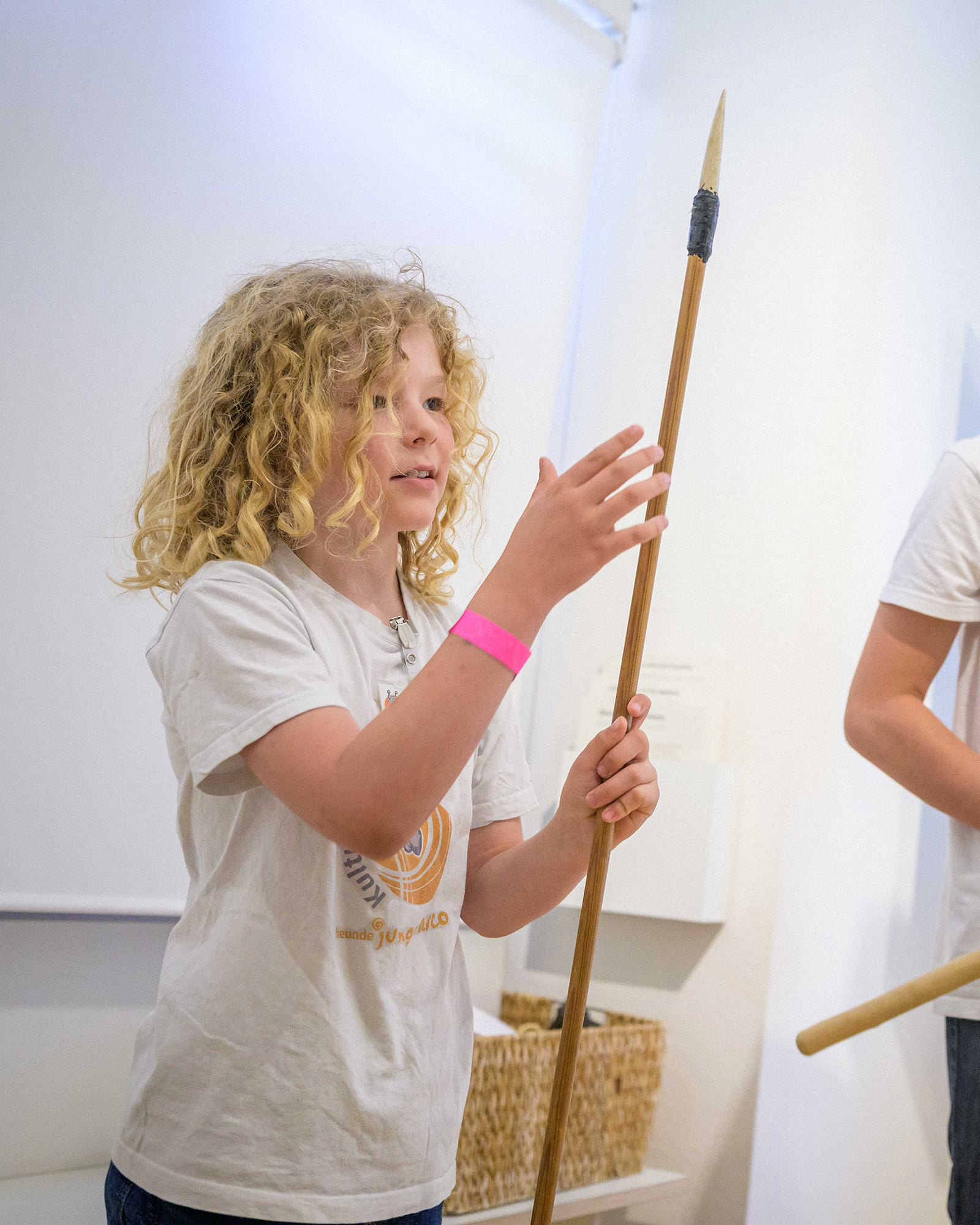 Ein Kind mit lockigem Haar hält einen langen Holzspeer mit dunkler Spitze und berührt ihn mit der anderen Hand. Im Hintergrund ist teilweise eine weitere Person mit einem Holzstab zu sehen. (KI-Alternativtext)