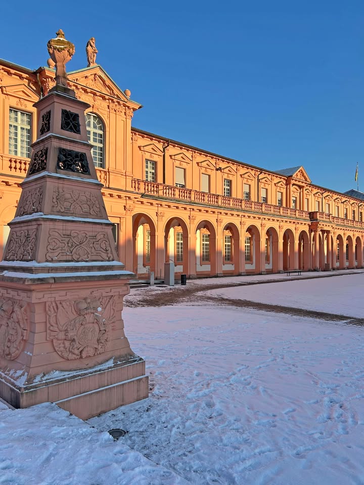Dieser Alternativtext wurde KI-generiert: Ein Gebäude mit einer Fassade aus Säulen und Bögen steht im Schnee. Das Licht der Sonne scheint auf die rosa-farbene Struktur.