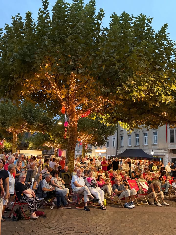 Eine große Menschenmenge sitzt auf roten Klappstühlen auf einem gepflasterten Platz und blickt in eine Richtung. Große Bäume im Hintergrund sind mit warmen Lichterketten geschmückt. Auf der Rückenlehne eines Stuhls ist der Schriftzug „Roter Hahn“ zu erkennen. (KI-Alternativtext)