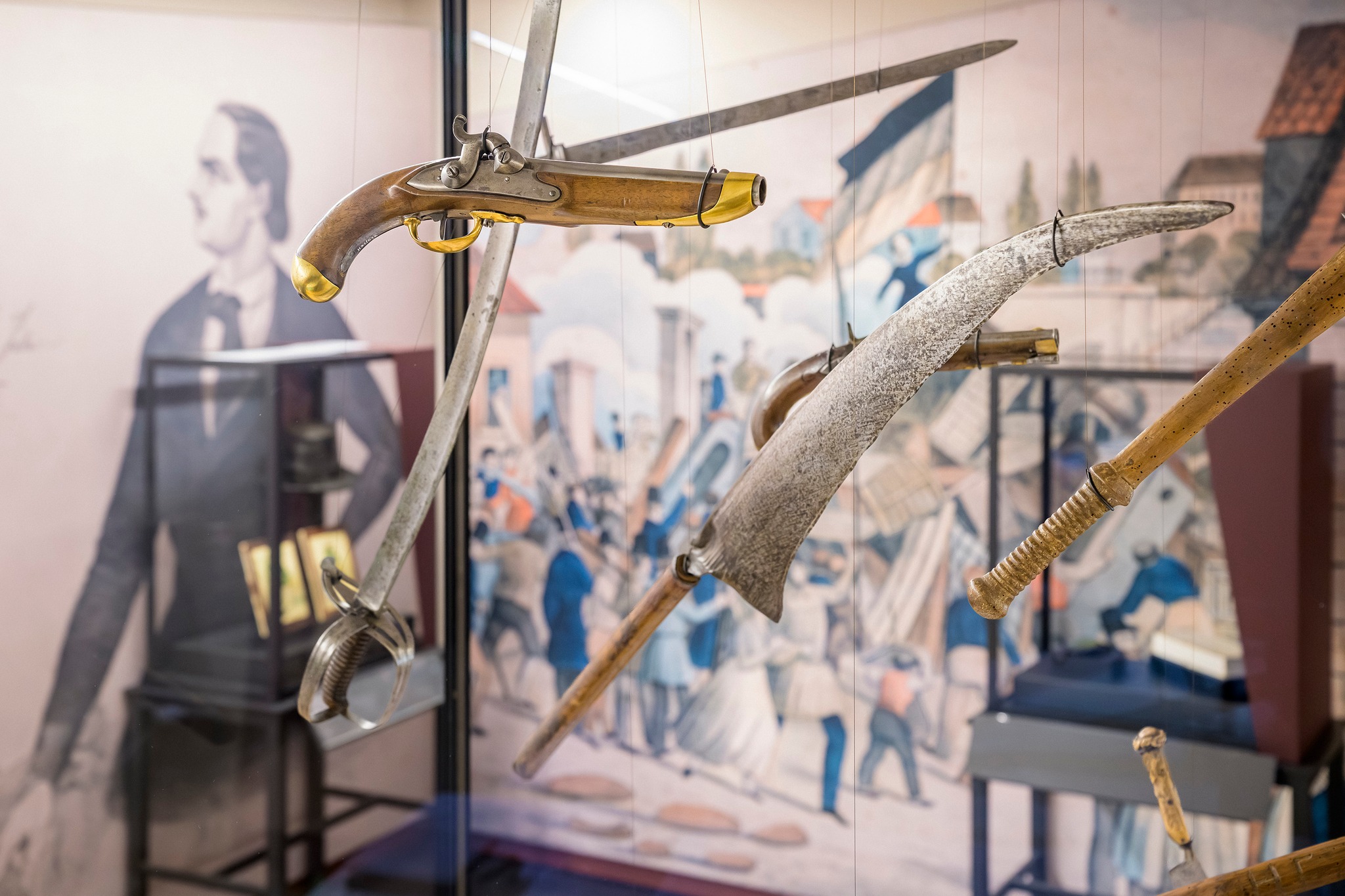 Historische Waffen wie Pistolen, Säbel und eine Sense hängen in einer Vitrine vor einer Wandmalerei, die eine Menschenmenge darstellt. Im Hintergrund ist eine große Schwarz-Weiß-Fotografie einer Person zu sehen. (KI-Alternativtext)