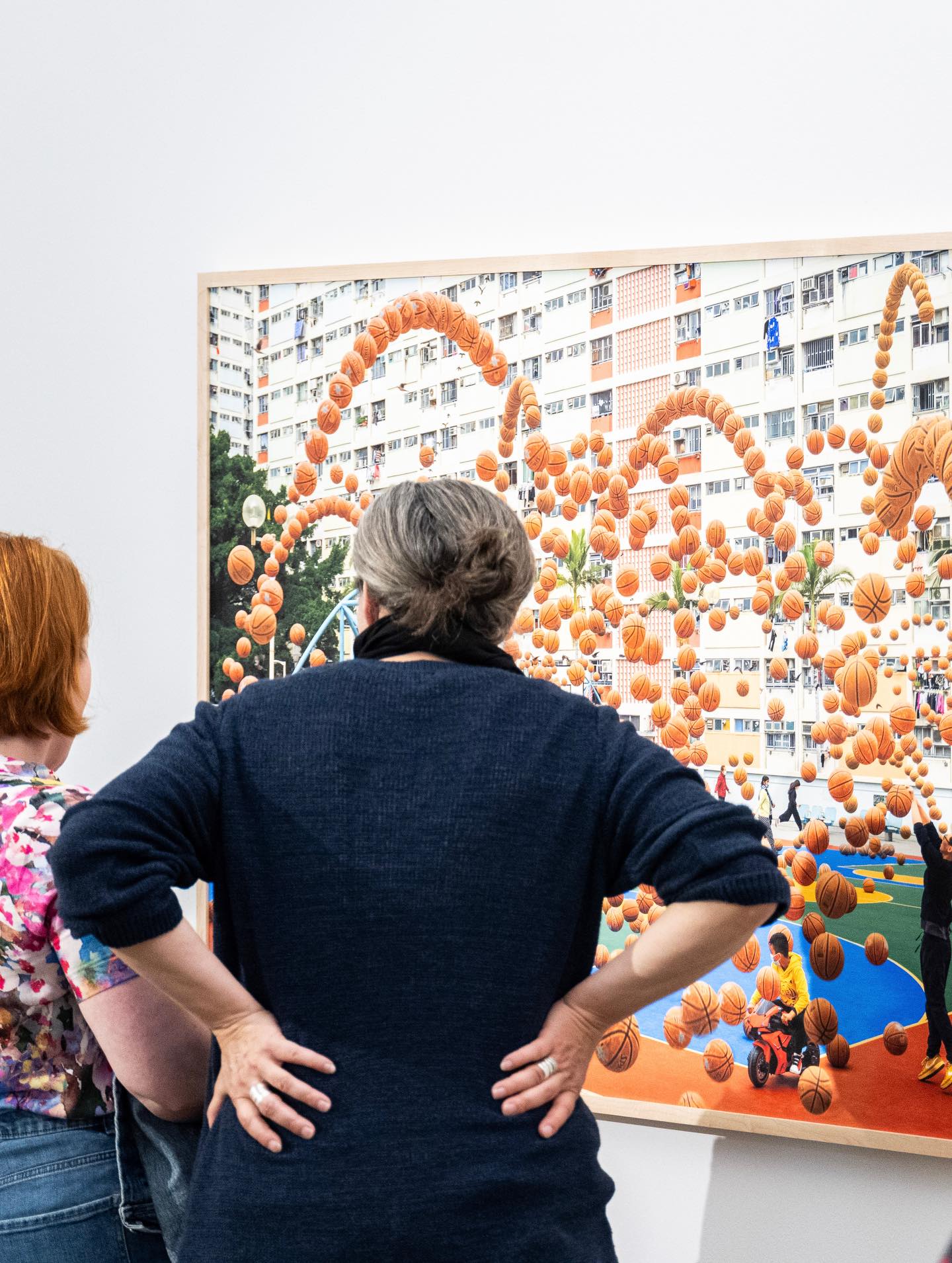 Dieser Alternativtext wurde KI-generiert: Zwei Personen betrachten ein farbenfrohes, großformatiges Kunstwerk an einer Gebäudewand. Das Kunstwerk zeigt Basketballbälle vor der Kulisse eines Wohnhauses.
