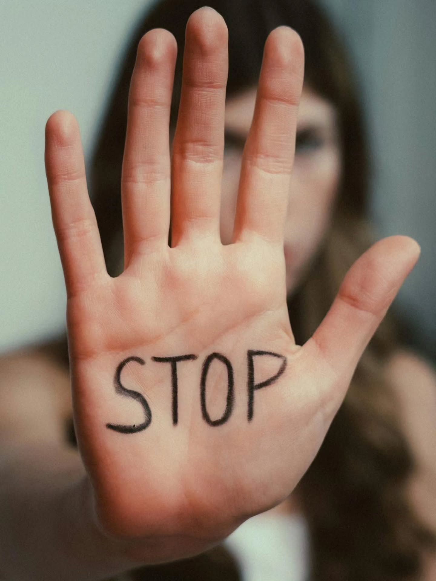 Eine Hand ist nach vorne gestreckt, auf der Handfläche steht in schwarzer Schrift das Wort "STOP". Im Hintergrund ist eine Person unscharf zu erkennen. (KI-Alternativtext)