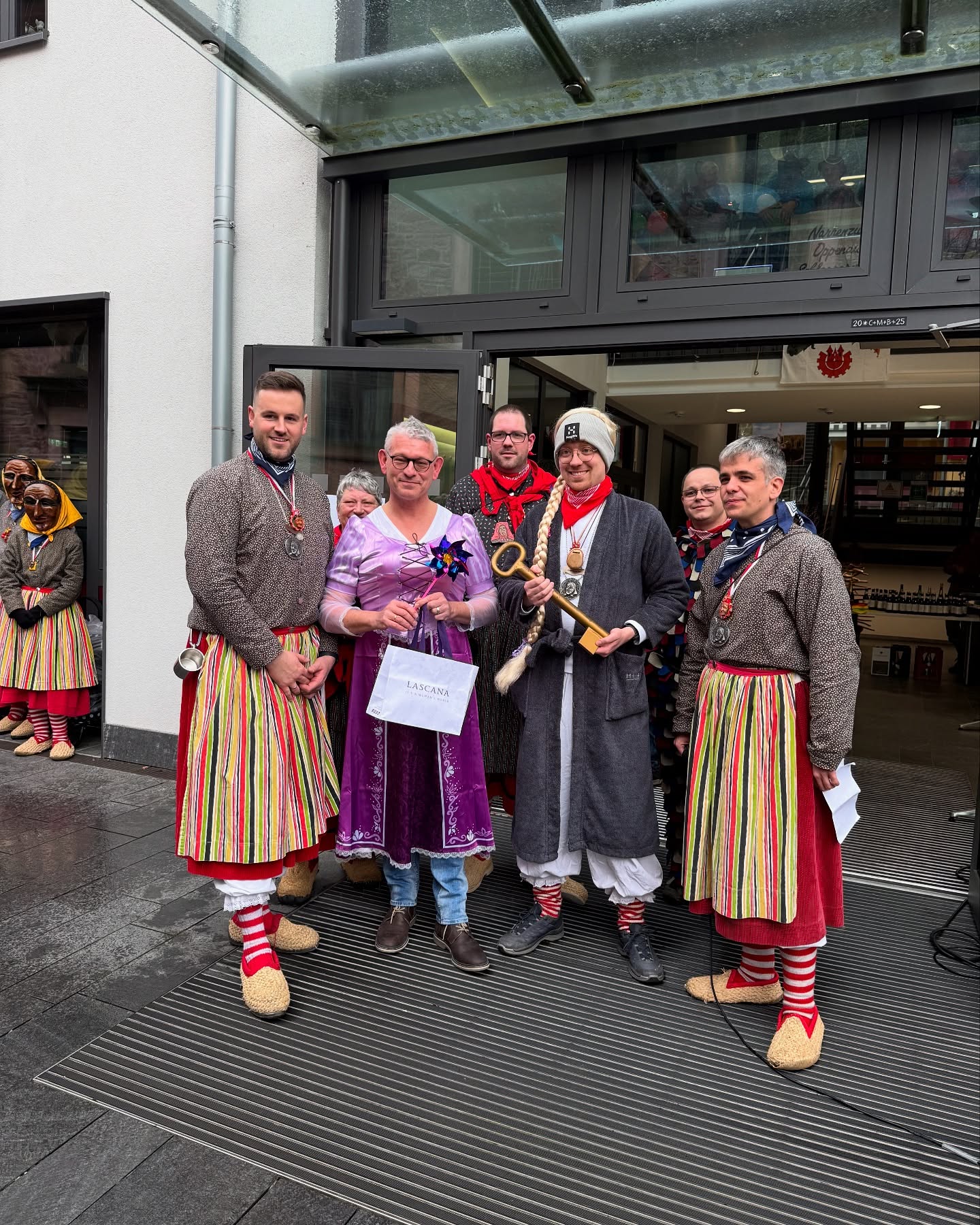 Eine Gruppe von Personen in unterschiedlichen Kostümen steht vor einem Geb"aude. Einige halten Gegenstände wie einen großen goldenen Schl"ussel und eine Tasche.