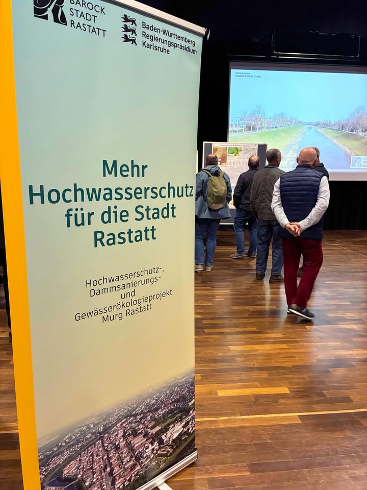 Dieser Alternativtext wurde KI-generiert: Ein farbiges Banner mit Text und Bild steht im Innenraum. Mehrere Personen stehen davor und betrachten ein Informationsmaterial.