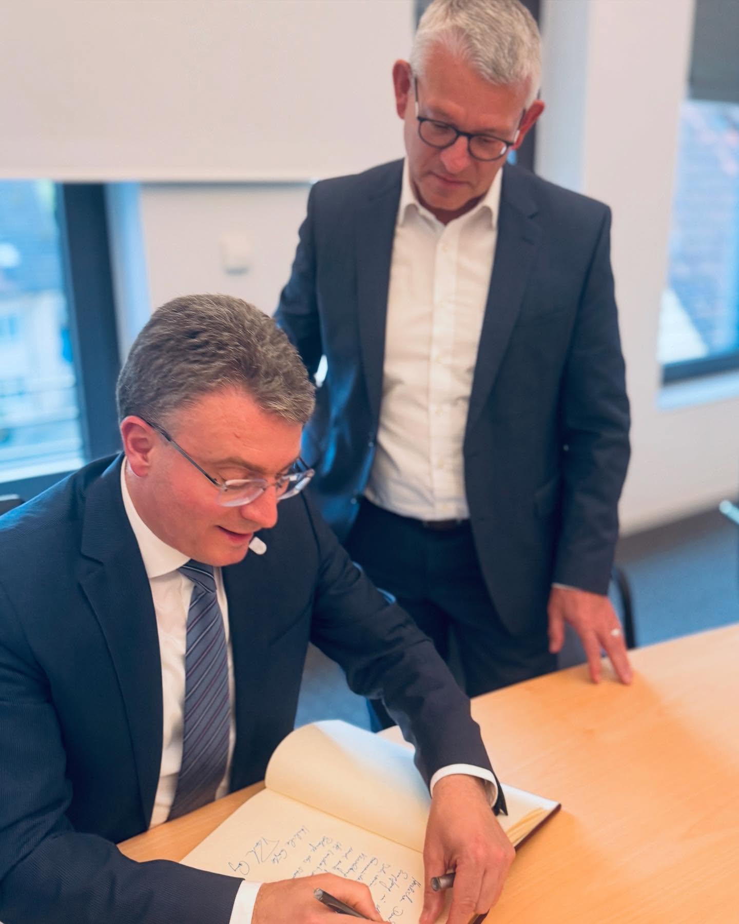 Zwei Personen in Anzügen an einem Tisch. Eine Person unterschreibt in ein Buch, während die andere Person zusieht.