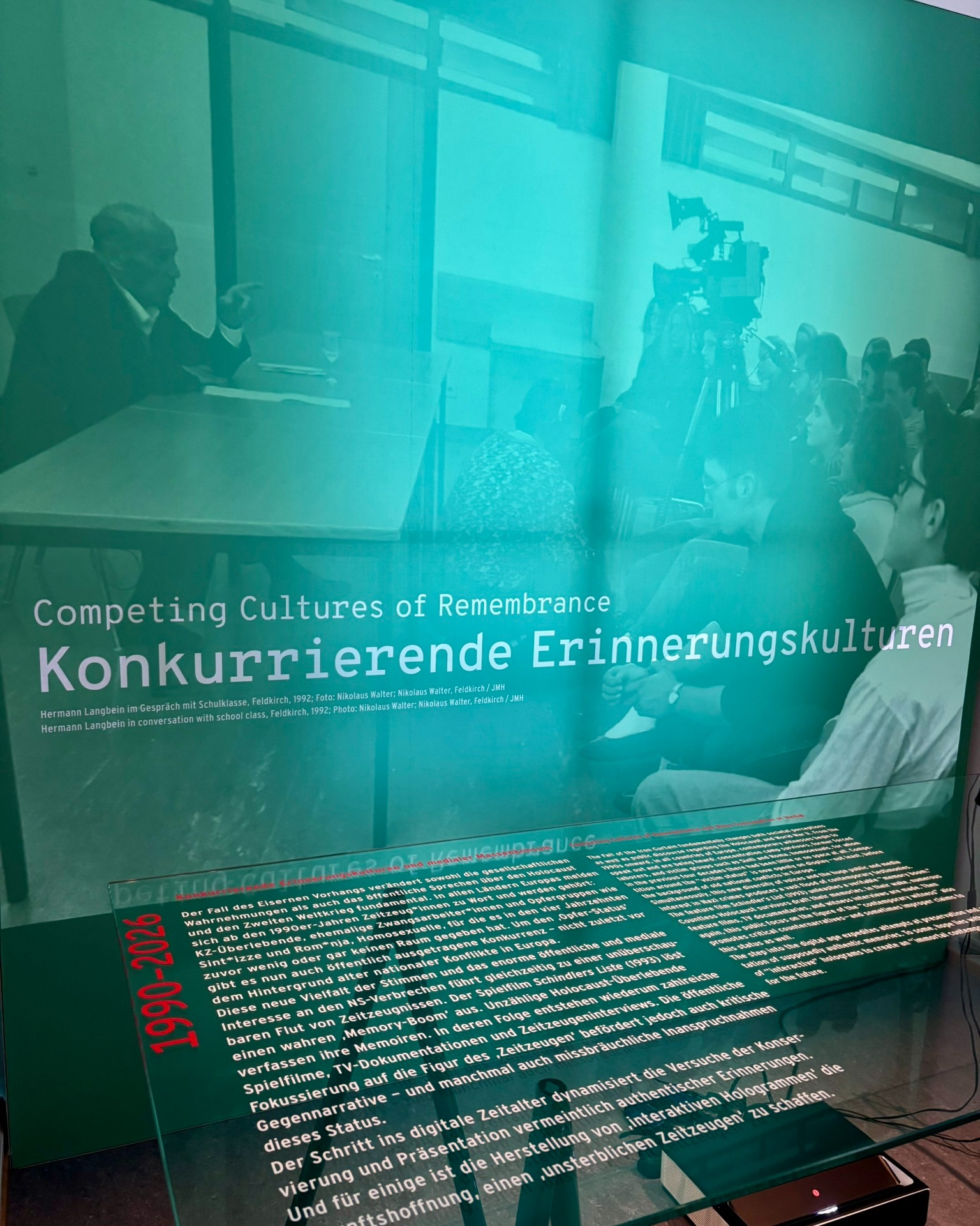 Ein Museumsdisplay mit der Überschrift „Competing Cultures of Remembrance / Konkurrierende Erinnerungskulturen“. Im Hintergrund sitzt eine Person an einem Tisch und spricht mit einer Gruppe von Zuhörern, während eine Kamera die Szene aufnimmt. Im Vordergrund liegt eine Glasscheibe mit einem mehrsprachigen Textblock, der den Zeitraum 1990-2026 und die Entwicklung der Erinnerungskultur thematisiert. (KI-Alternativtext)