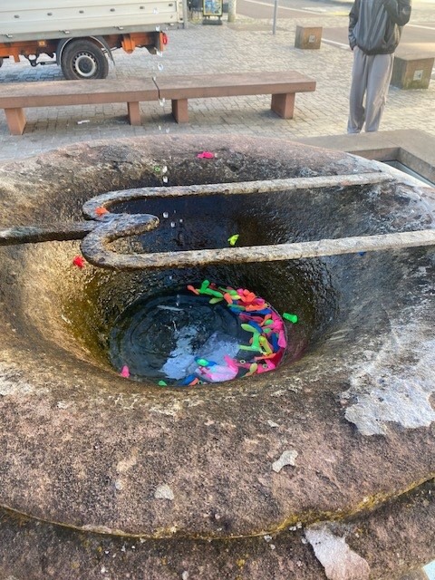 Steinbrunnen mit Wasser und bunten Plastikfiguren am Boden, darüber ein Metallgriff. Im Hintergrund Holzbank, LKW und stehende Person. (KI-Alternativtext)
