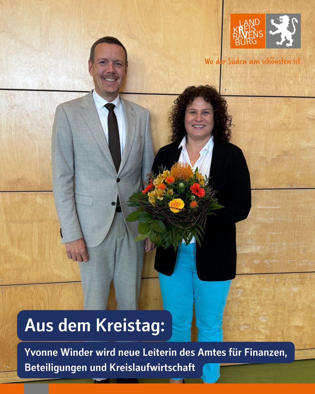 Dieser Alternativtext wurde KI-generiert: Zwei Personen stehen nebeneinander vor einer hellen Wand. Eine Person hält einen Blumenstrauß, ein Logo und Text befinden sich im Bild.