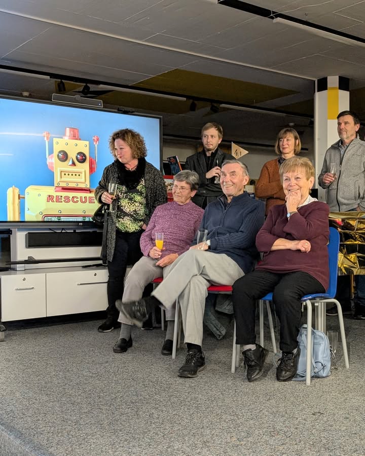 Dieser Alternativtext wurde KI-generiert: Eine Gruppe von Personen sitzt und steht in einem Innenraum vor einer Leinwand mit einer Roboterabbildung. Einige halten Gläser oder Becher in der Hand.