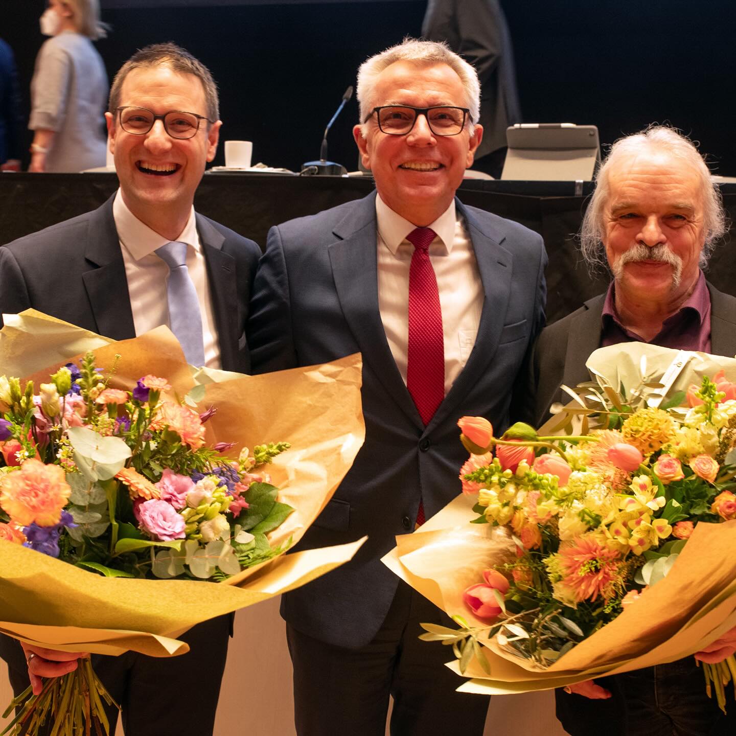 Dieser Alternativtext wurde KI-generiert: Drei Personen in Anzügen lächeln in die Kamera und halten Blumensträuße. Der Hintergrund zeigt einen Raum mit einem Pult und weiteren Personen.