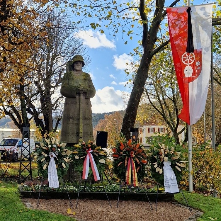 Dieser Alternativtext wurde KI-generiert: Eine Statue steht vor Bäumen und einem Gebäude. Die Statue ist mit Blumen und Wimpelketten geschmückt.