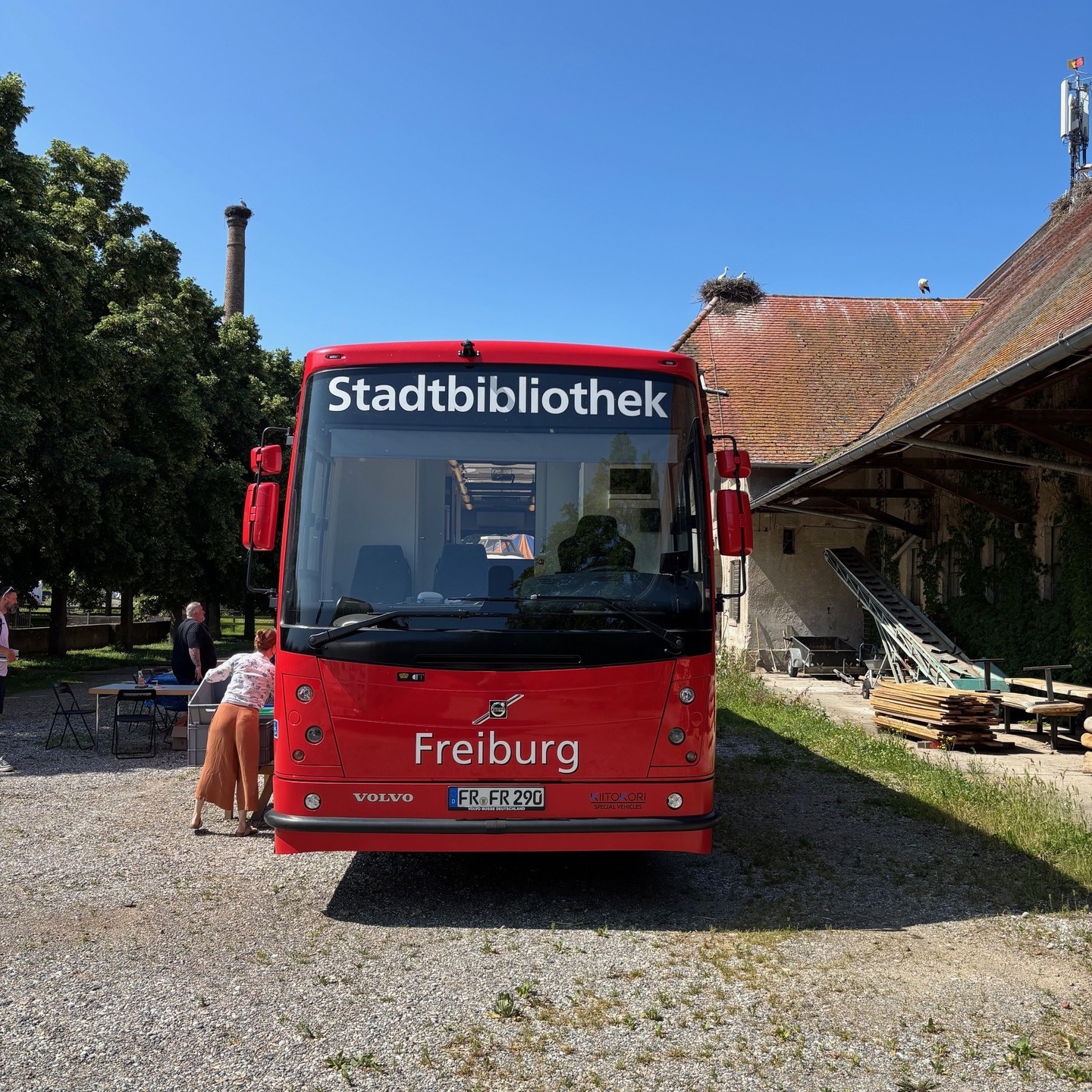 Ein roter Bus mit der Aufschrift "Stadtbibliothek Freiburg" steht vor einem Gebäude mit einem Storchennest auf dem Dach. Eine Person steht in der Nähe des Busses.