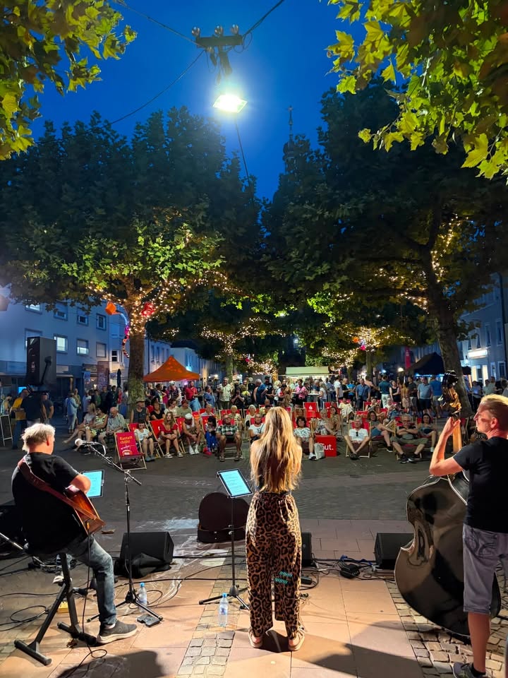 Eine Band spielt auf einer kleinen Bühne im Freien bei Dämmerung. Links sitzt eine Person mit Gitarre, in der Mitte steht eine Person in gemusterten Hosen am Mikrofon und rechts steht eine Person mit Kontrabass. Davor sitzt ein Publikum auf roten Stühlen unter Bäumen mit Lichterketten, wobei rote Schilder mit der Aufschrift „Kultur im Park“ zu sehen sind. (KI-Alternativtext)