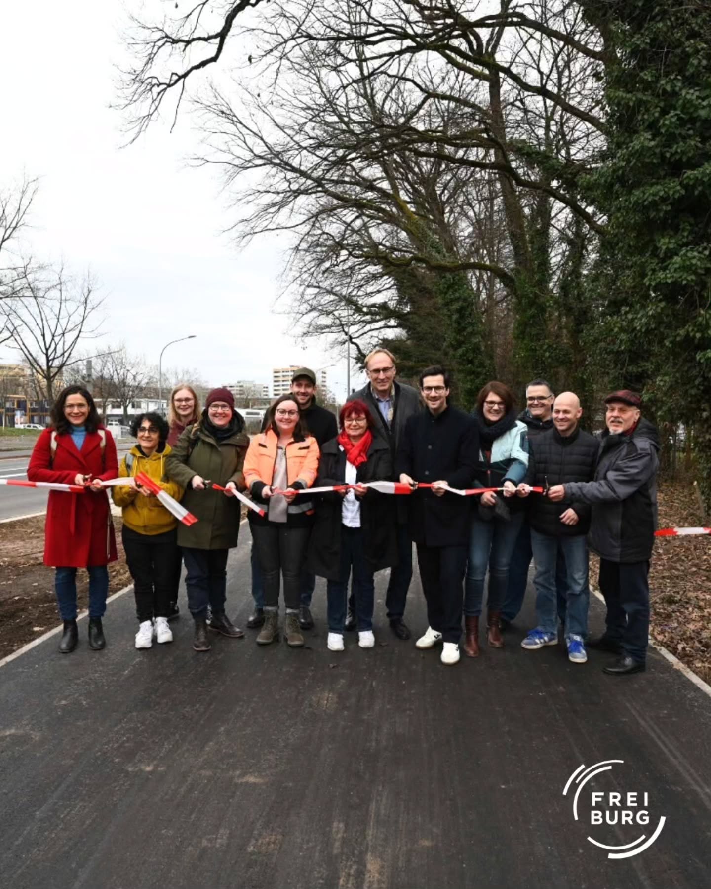 Dieser Alternativtext wurde KI-generiert: Eine Gruppe von Personen steht auf einem neuen oder renovierten Gehweg und hält rote Bänder. Im Hintergrund sind Bäume und Gebäude zu sehen.