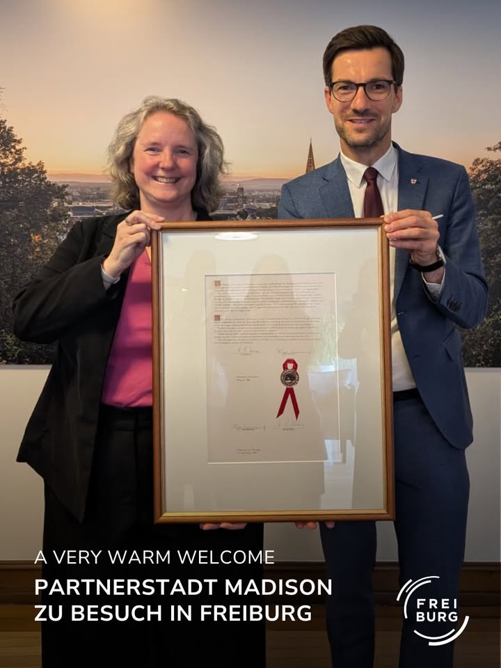 Zwei Personen halten gemeinsam ein gerahmtes Dokument mit rotem Band und Siegel. Im Hintergrund eine Stadtansicht bei Sonnenuntergang. Unten steht: „A VERY WARM WELCOME PARTNERSTADT MADISON ZU BESUCH IN FREIBURG“ und das Logo „FREIBURG“. (KI-Alternativtext)