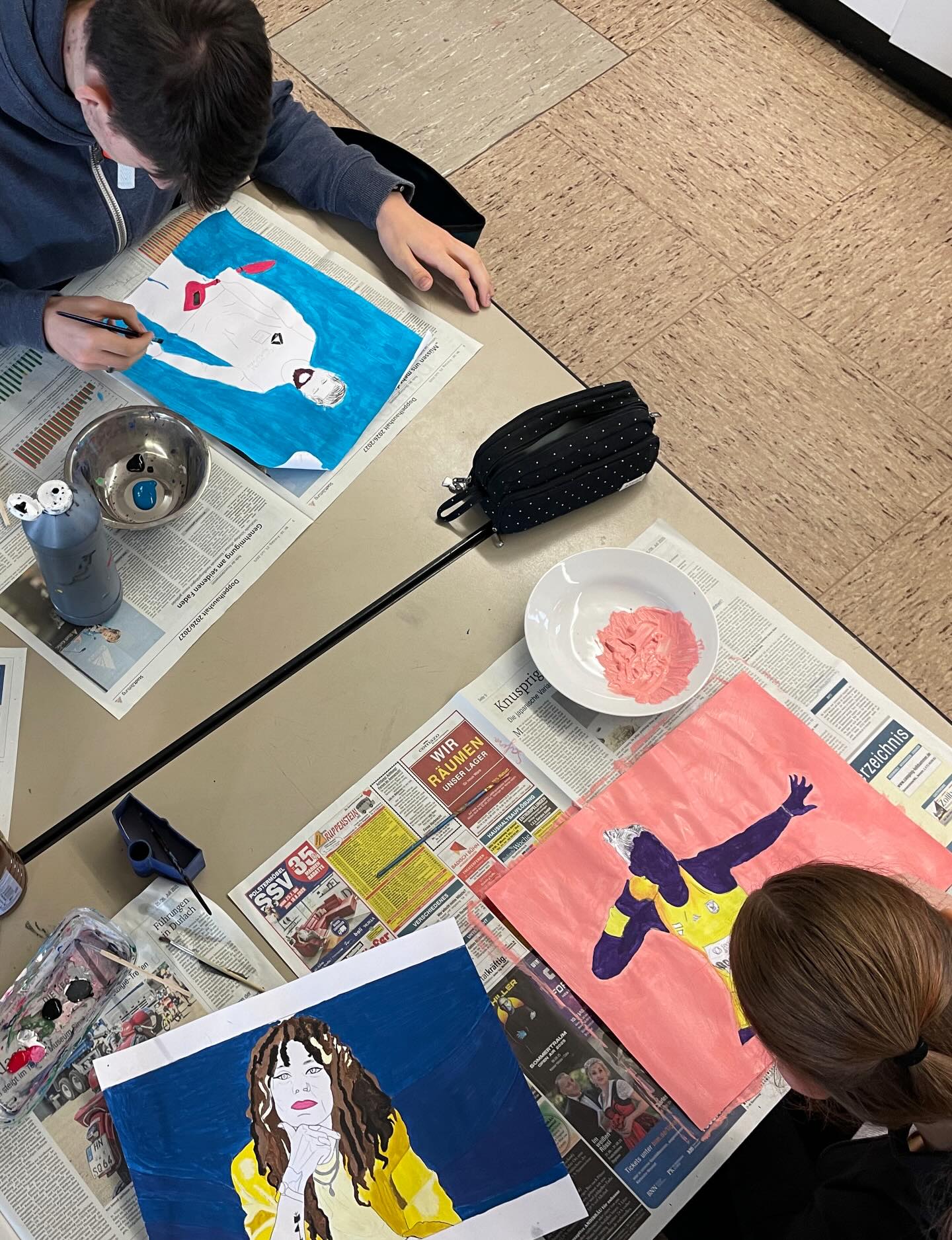 Dieser Alternativtext wurde KI-generiert: Ein Tisch ist mit Zeitungspapier bedeckt und zeigt verschiedene Kunstwerke in Bearbeitung. Es sind Farbtöpfe und Pinsel zu sehen.