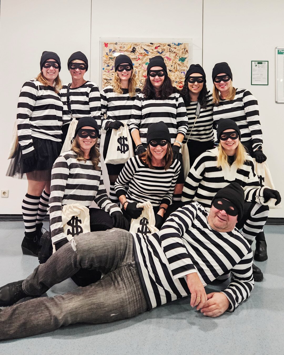 Dieser Alternativtext wurde KI-generiert: Eine Gruppe von Personen trägt gestreifte Kleidung und Masken. Einige halten Geldsäcke, während eine Person am Boden liegt.