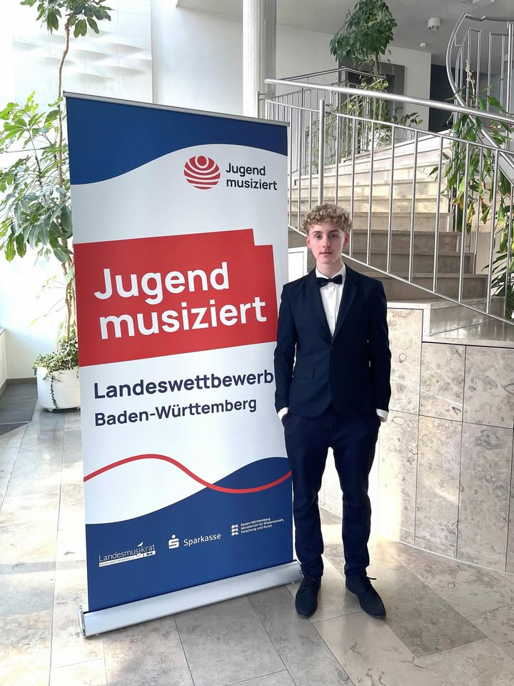 Dieser Alternativtext wurde KI-generiert: Eine Person in formeller Kleidung steht vor einem Werbebanner. Das Banner ist weiß mit rotem Text und Logos.