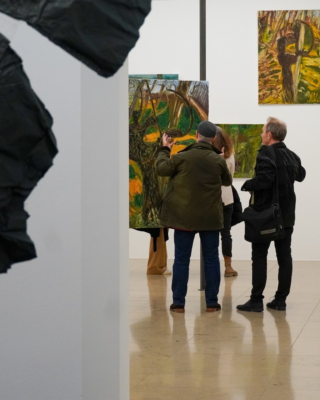 Dieser Alternativtext wurde KI-generiert: Innenraum einer Kunstausstellung mit mehreren Gemälden an den Wänden. Besucherinnen und Besucher betrachten ein Bild und diskutieren darüber.