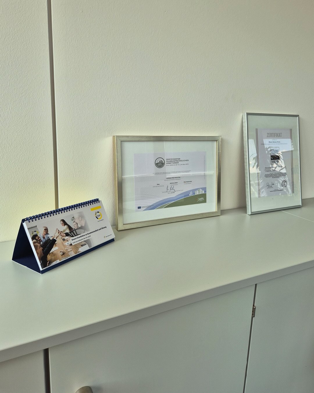 Auf einer weißen Ablagefläche stehen von links nach rechts ein Tischkalender mit einer Gruppenfotografie, ein gerahmtes Zertifikat mit der Aufschrift „Zertifizierter fahrradfreundlicher Arbeitgeber“ und ein weiteres gerahmtes Zertifikat. (KI-Alternativtext)