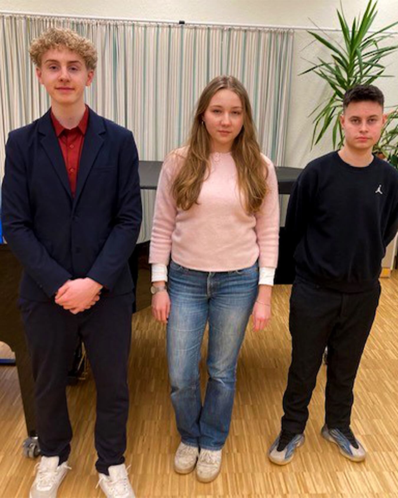 Dieser Alternativtext wurde KI-generiert: Drei Personen stehen nebeneinander vor einem Hintergrund mit Vorhängen und Pflanzen. Sie tragen formelle und lässige Kleidung.