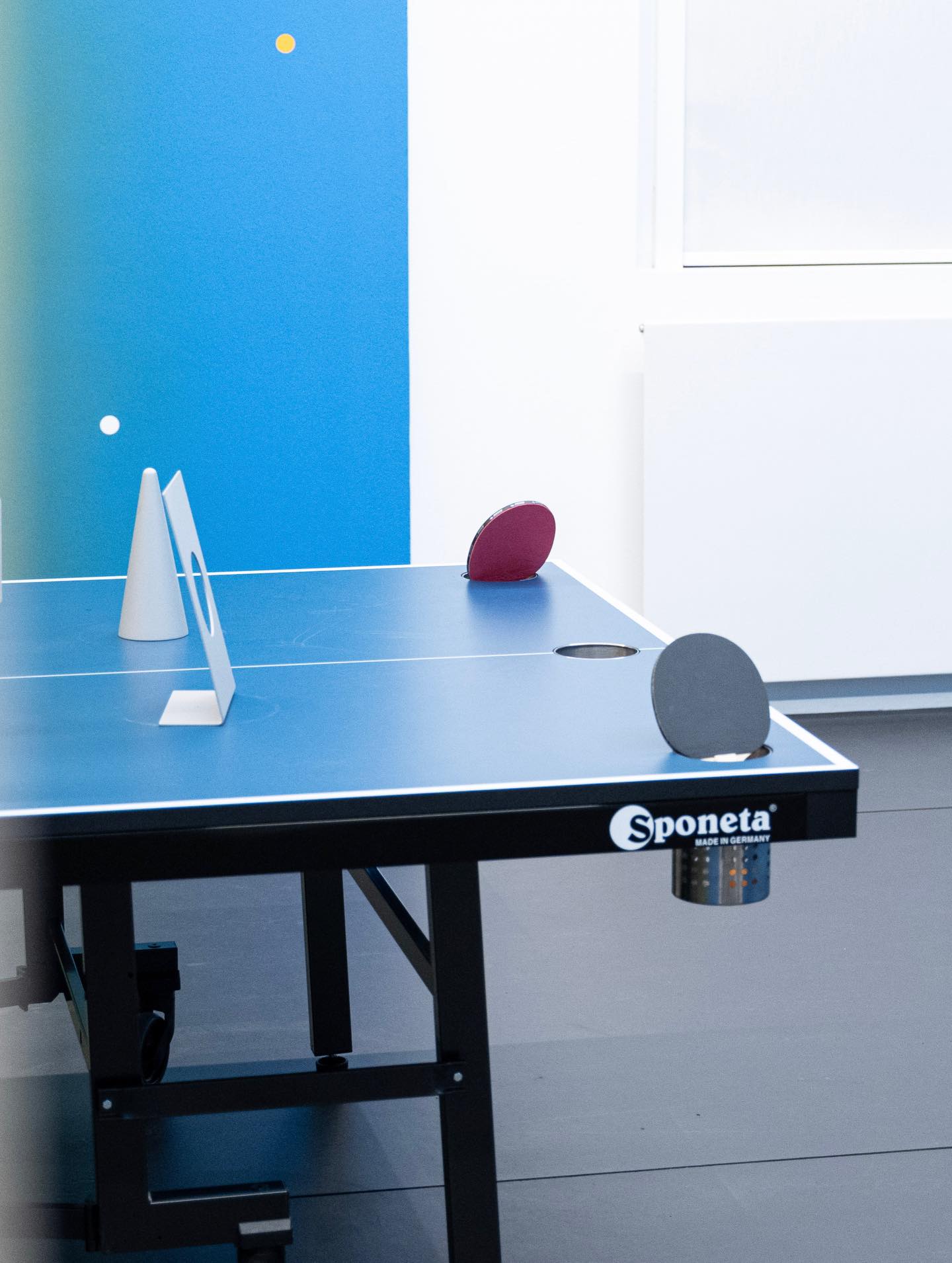Dieser Alternativtext wurde KI-generiert: Ein Tischtennistisch mit Zubehör, darunter Bälle und Schläger. Der Hintergrund ist eine blaue Wand und ein helles Zimmer.