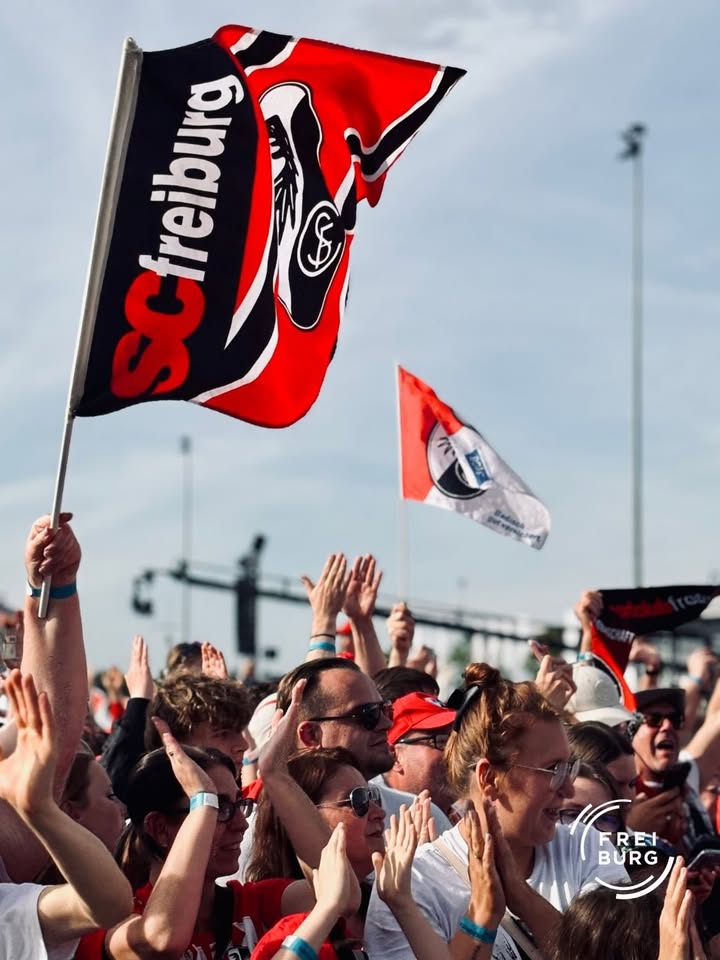 Eine Menschenmenge hebt die Hände und schwenkt eine große rote und schwarze Fahne mit der Aufschrift „SC Freiburg“ und dem Vereinslogo. Im Hintergrund ist eine weitere, kleinere Fahne sowie ein Stadionzaun zu erkennen. (KI-Alternativtext)
