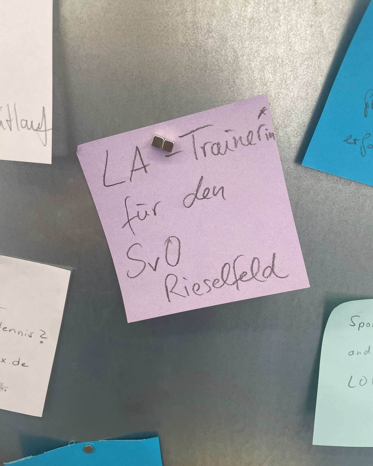 Eine rosa Haftnotiz ist mit einem kleinen Metallclip an einer grauen Oberfläche befestigt. Darauf steht handschriftlich: „LA = Trainerin für den SvO Rieselfeld“. Um die Notiz herum sind Teile weiterer Zettel sichtbar. (KI-Alternativtext)