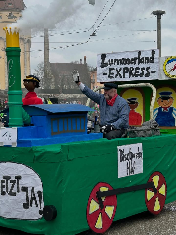 Dieser Alternativtext wurde KI-generiert: Ein farbenfrohes, selbstgebautes Gefährt im Zugstil ist mit Figuren und Aufschriften dekoriert. Im Hintergrund sind Gebäude und Bäume zu sehen.