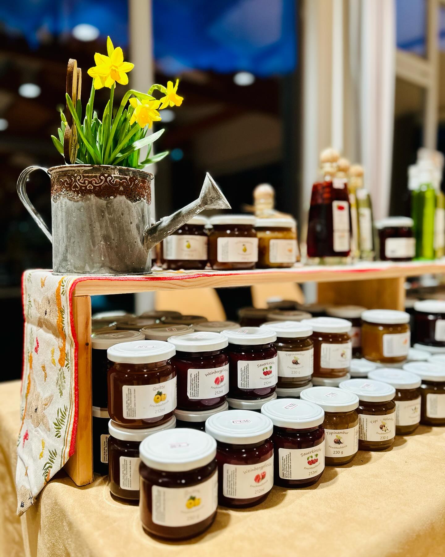 Blumen in einer Giesskanne stehen auf einem Regal. Regalböden sind voll mit Gläsern, auf denen Etiketten kleben.