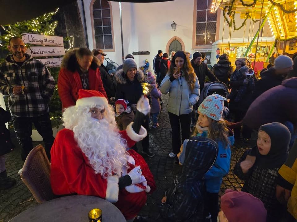 Dieser Alternativtext wurde KI-generiert: Eine Gruppe von Menschen steht um jemanden in einem roten Anzug mit weißem Bart. Im Hintergrund befindet sich ein beleuchteter Weihnachtsmarktstand und ein geschmückter Baum.