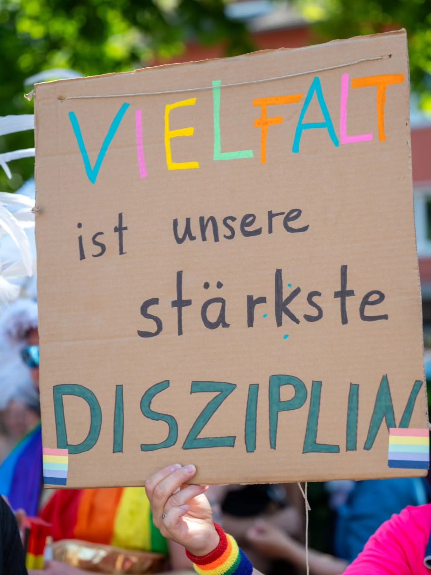 Ein Kartonschild mit der Aufschrift „VIELFALT ist unsere stärkste DISZIPLIN“ in bunten Buchstaben, unten mit zwei Regenbogenflaggen verziert, wird von einer Hand gehalten. Im Hintergrund sind unscharf Menschen und grüne Bäume zu erkennen. (KI-Alternativtext)