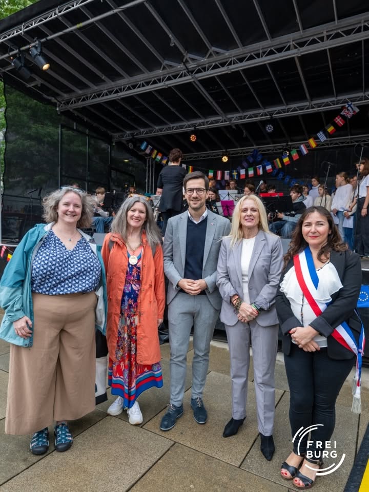 Fünf Personen stehen vor einer Bühne mit Flaggenketten und einem Orchester im Hintergrund. Eine trägt eine rot-weiß-blaue Amtsschärpe, alle lächeln in die Kamera. Unten rechts ist das Logo „FREIBURG“ sichtbar. (KI-Alternativtext)