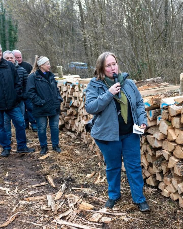 Dieser Alternativtext wurde KI-generiert: Mehrere Personen stehen im Freien neben einem Holzstapel. Eine Person hält ein Mikrofon und spricht, während andere zuhören.